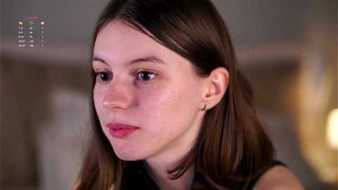 Media: Video of a young Caucasian woman with fair skin, brown eyes, and straight brown hair, wearing a black top, looking contemplative. Background is blurred with soft lighting.