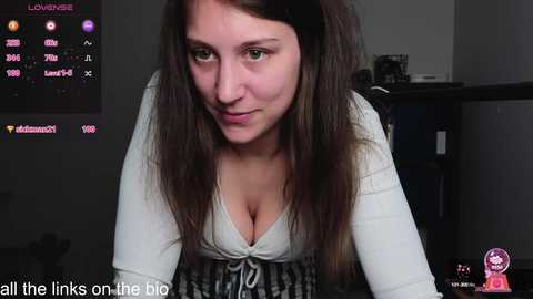 Media: A video of a young woman with long brown hair, wearing a white top, leaning forward in a dimly lit room with a TV screen showing her stats.