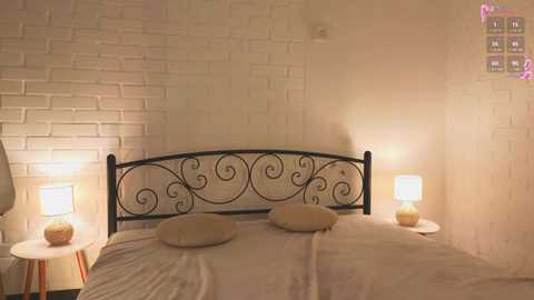 Media: Video of a cozy bedroom with a black wrought-iron headboard, beige brick wall, two bedside lamps with white shades, and a neatly made bed with beige linens.