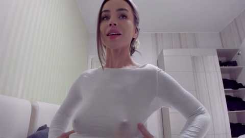 Media: Video of a fit woman with dark hair, wearing a tight white long-sleeve top, standing confidently in a modern, white-tiled bathroom with cabinets and a shower.