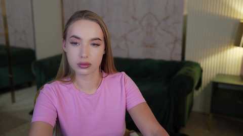 Media: Video of a young woman with fair skin, long blonde hair, and a pink t-shirt, looking introspective, sitting indoors with green couches, patterned wallpaper, and a lit lamp in the background.