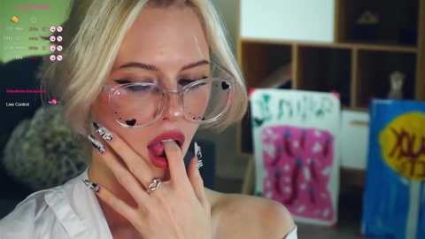 Media: A video of a blonde woman with fair skin, wearing clear glasses and a white shirt, seductively biting her finger while sitting in a room with a pink towel and a soda can.