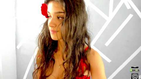 Media: Video of a young woman with long, wavy brown hair adorned with a red rose, wearing a red lace bralette, against a white geometric background.