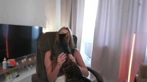 Media: A video captures a woman with long, wavy hair, wearing a sleeveless top, seated in a dark chair, petting a black and white cat. The background features a flat-screen TV, curtains, and a floor lamp, giving a cozy, indoor setting.