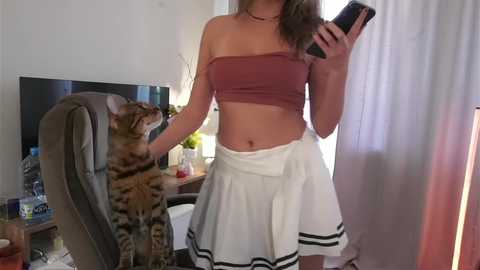 Media: Video of a woman in a maroon bandeau top and white pleated skirt, holding a phone, standing next to a striped tabby cat in a living room with a TV, chair, and windows.