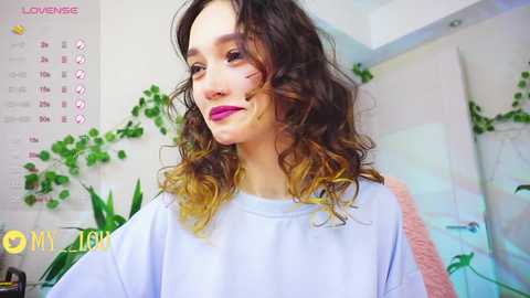 Media: Video of a young woman with curly, multi-colored hair in a white t-shirt, smiling, in a bright room with plants and a calendar.