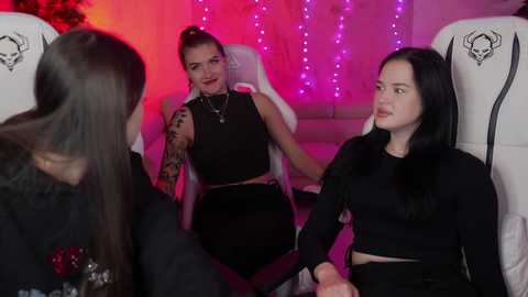 Media: Video of three women with long black hair, sitting in white recliner chairs, holding hands, in a dimly-lit room with pink and purple LED lights.