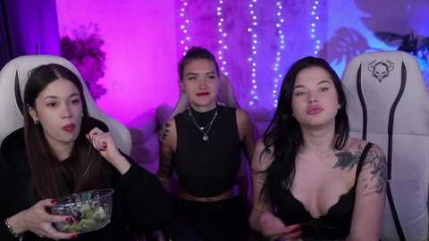 Media: Video of three women in black outfits, one with a tattoo sleeve, sitting in gaming chairs, illuminated by purple and blue LED lights.