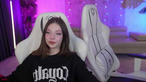 Media: Video of a young woman with fair skin and straight brown hair, wearing a black \"Riot Games\" hoodie, seated in a white gaming chair under purple and blue LED lighting in a modern, dimly lit room.