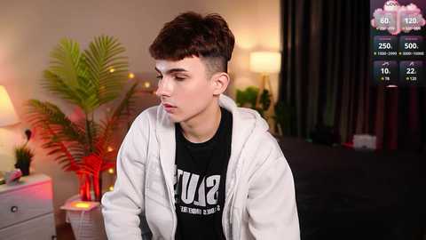 Media: Video of a young man with short, dark hair and a shaved side, wearing a white hoodie over a black T-shirt, in a dimly lit bedroom with a bed, plants, and digital fitness tracker overlay.