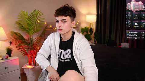 Media: Video of a young man with short brown hair, wearing a white hoodie over a black shirt, sitting on a bed in a dimly-lit room with potted plants, a TV, and a nightstand.