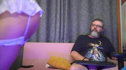 Media: Video of a man with a beard and glasses sitting on a couch, wearing a black T-shirt, looking at a woman in white lingerie in the foreground.