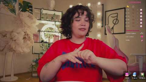 Media: Video of a plus-size woman with curly black hair, wearing a red t-shirt with \"BRAVE\" written in blue, sitting in a cozy room with fairy lights and art on the wall.