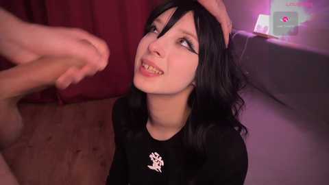 Media: A video of a young woman with long black hair, fair skin, and dark eyeliner, kneeling on a wooden floor. She wears a black shirt with a white emblem. Her expression is playful as she is touched by another person's hand.