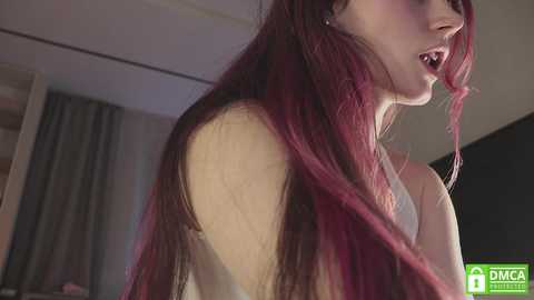 Media: Video of a fair-skinned woman with long, straight, deep red hair, wearing a white top, her mouth open as if in mid-speech, in a dimly-lit indoor setting with a blurred background.