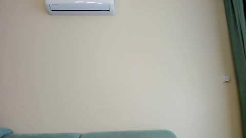 Media: Video of a minimalist room with a beige wall, a white air conditioning unit above, a green curtain on the right, and a light blue sofa at the bottom.