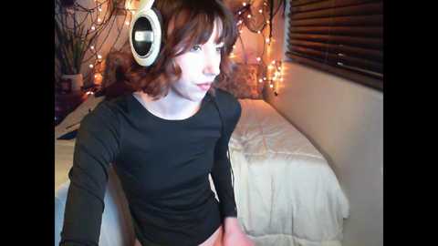 Media: Video of a slender, pale-skinned woman with short, wavy brown hair, wearing a black long-sleeve top and large white headphones, sitting on a bed with white bedding, surrounded by warm, string lights and dark wooden blinds in a cozy bedroom.