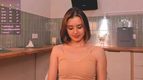 Media: Video of a young woman with light skin and brown hair in a beige camisole, sitting in a modern kitchen with green tiled walls, wooden countertops, and a black flat-screen TV above.