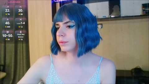 Media: A video of a young woman with short, vibrant blue hair, wearing a white lace bralette, standing indoors with a wooden background.