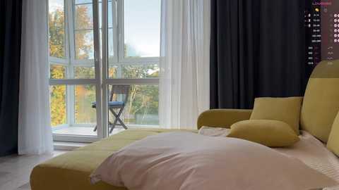 Media: Video of a modern bedroom with a large bed covered in yellow and white linens, dark curtains, and a glass wall showcasing a patio with a wooden chair and greenery outside.