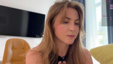 Media: Video of a fair-skinned woman with long, light brown hair, wearing a black top, sitting indoors near a large TV and yellow chair.