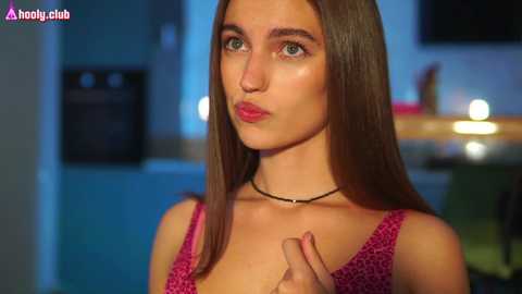 Media: Video of a young woman with straight brown hair, light skin, and green eyes, wearing a pink patterned top and black necklace, in a dimly lit room with blurred furniture.