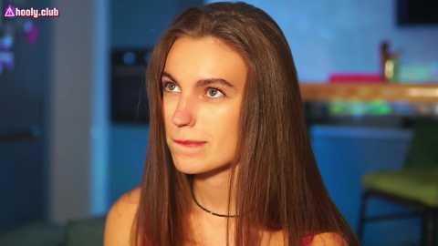 Media: Video of a fair-skinned, long-haired brunette woman with green eyes, wearing a red top, in a modern, dimly lit living room with blurred background details.