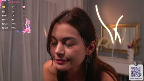 Media: Video of a smiling, light-skinned Latina woman with dark hair, wearing black earrings, in a modern, dimly lit room with a large mirror, floral decor, and digital overlays.