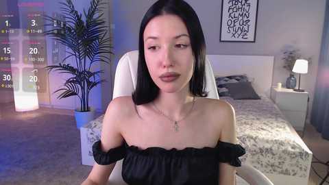 Media: Video of a young woman with long black hair, wearing a black off-shoulder dress, sitting on a white chair in a modern bedroom with a potted plant, white bed, and abstract artwork.