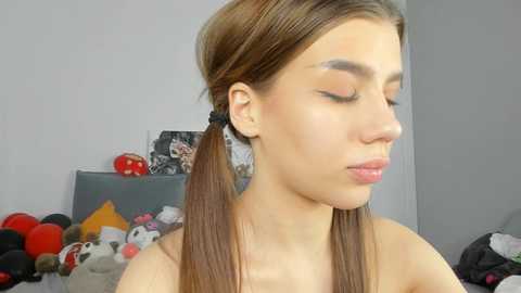 Media: A video of a young, light-skinned woman with straight brown hair styled in pigtails, wearing black scrunchies. She has a neutral expression and is indoors, with a cluttered background featuring stuffed animals and a gray wall.