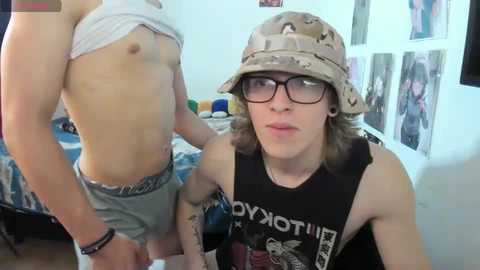 Media: Video of a muscular man with light skin, wearing a camouflage bucket hat, lifting his shirt to reveal his toned stomach, and a young woman with glasses and a black tank top.