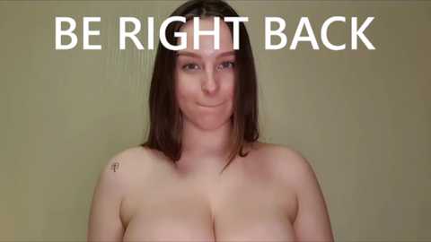 Media: Video of a smiling, topless woman with large breasts, fair skin, and straight brown hair, against a plain, olive-green background. Text above reads, \"BE RIGHT BACK.\