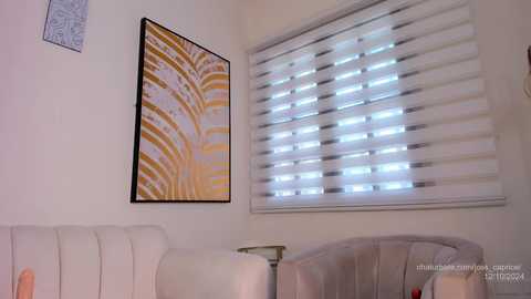 Media: Video of a modern living room with a beige sofa, white blinds covering a window, and a framed abstract painting with yellow and white patterns.