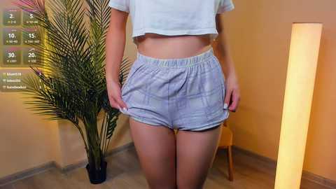 Media: Video of a light-skinned woman wearing a white crop top and light blue plaid shorts standing in a room with a tall potted palm plant and a lit lamp.