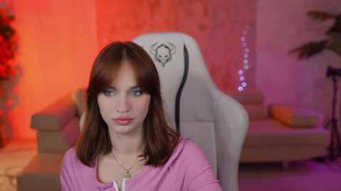 Media: A video of a fair-skinned woman with shoulder-length brown hair, wearing a pink shirt, sitting in a white gaming chair, surrounded by colorful LED lights and a red flower arrangement.