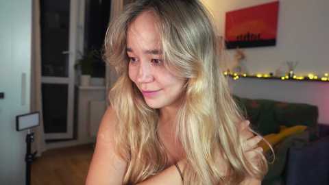 Media: Video of a blonde, fair-skinned woman with long, wavy hair, wearing a pink top, sitting on a sofa in a cozy living room with a wooden floor, illuminated by warm lighting.