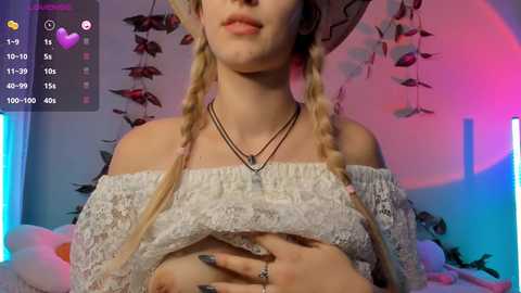 Media: Video of a fair-skinned woman with blonde braided hair, wearing an off-shoulder lace top, a black necklace, and a hat. She stands in a room with purple and blue lighting, and decorative plants.