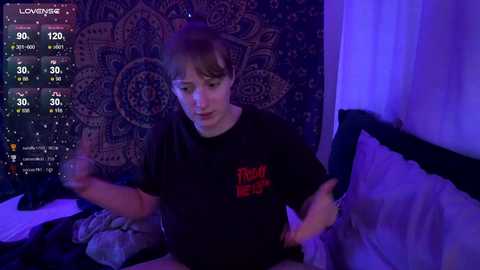 Media: Video of a young person with light skin, short brown hair, wearing a black t-shirt with red text, sitting on a bed with a dark patterned tapestry background, under blue lighting.