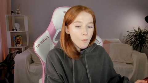 Media: Video of a young redhead woman with fair skin, wearing a gray hoodie, sitting in a pink and white gaming chair, in a modern living room with beige couches and a potted plant.