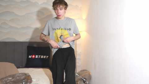 Media: A video shows a young, slender Caucasian male with short, tousled brown hair, wearing a gray t-shirt with \"Dad\" and black pants, standing in a dimly lit bedroom with a modern, abstract wall mural and a bed with a \"NOSTREET\" pillow.