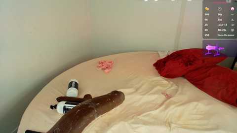 Media: Video of a messy bedroom with a large, realistic brown dildo on beige sheets, red pillow, scattered pink rose petals, and a white bottle of lubricant.