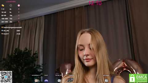 Media: Video of a young blonde woman with long hair, wearing a blue top, sitting in a room with brown curtains, recording herself on a streaming platform.