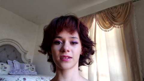 Media: Video of a young person with short, curly, dark hair, wearing a pink top, standing in a bedroom with a tufted headboard, floral-patterned bedspread, beige curtains, and soft lighting.