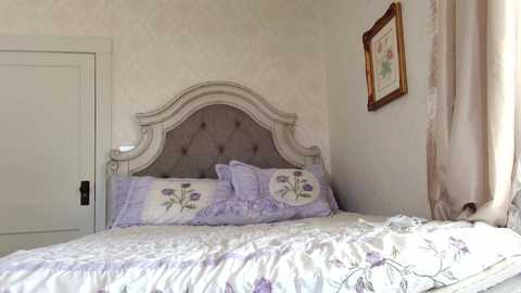 Media: Video of a serene bedroom featuring a queen-sized bed with a gray tufted headboard, lavender floral bedding, and framed artwork on a pale yellow wall.