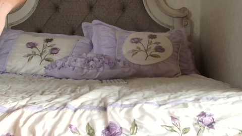 Media: Video of a cozy, pastel-colored bedroom featuring a tufted, upholstered headboard, lavender pillows, and a floral bedspread with purple roses. The room has a soft, feminine ambiance with a vintage, romantic style.