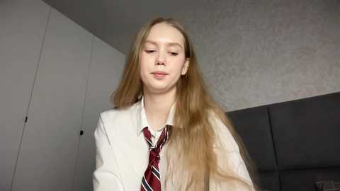 Media: Video of a young woman with long, straight blonde hair, wearing a white shirt and a red and white striped tie, standing indoors against a grey textured wall and white cabinets.