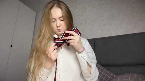 Media: Video of a young Caucasian girl with long, wavy blonde hair, wearing a white shirt and a red and black striped necktie. She is in a bedroom with a gray headboard and white walls.