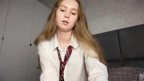 Media: Video of a young Caucasian woman with long blonde hair, wearing a white shirt and red tie, standing in a dimly lit bedroom with gray bedding and a gray headboard.