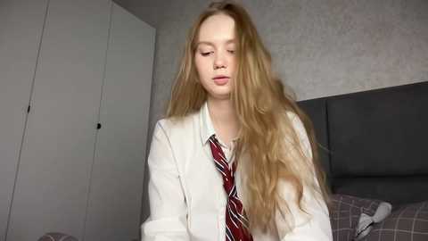 Media: Video of a fair-skinned, long-haired, blonde-haired young woman in a white shirt and red striped tie, standing in a modern bedroom with grey walls, a grey headboard, and a white wardrobe.