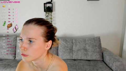 Media: Video of a young Caucasian woman with fair skin and light brown hair tied back, wearing a gold necklace, sitting on a gray sofa in a modern, minimally decorated room.